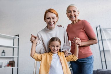 joyful lesbian couple holding hands of adopted daughter smiling at camera at home clipart