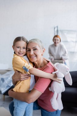 cheerful lesbian woman embracing adopted daughter and looking at camera in new home clipart