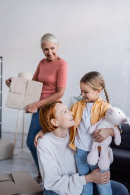 happy lesbian woman looking at adopted girl holding toy bunny in new apartment clipart