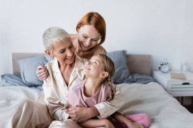 Woman hugging girlfriend in pajamas and child on bed  clipart