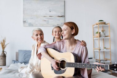 Gülümseyen kadın ve çocuk yatakta akustik gitar çalan kız arkadaşına bakıyor. 