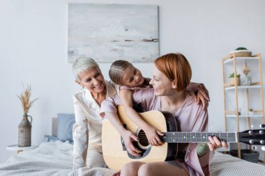 Yatakta evlatlık kız ve kız arkadaşının yanında akustik gitar çalan pozitif kadın. 
