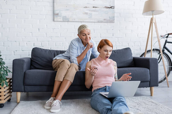 amazed woman showing wow gesture while sitting on floor with laptop near lesbian girlfriend on couch