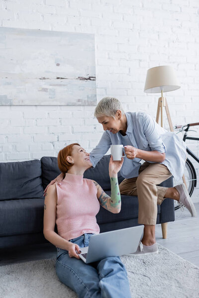 lesbian woman giving cup of tea to happy girlfriend sitting on couch with laptop