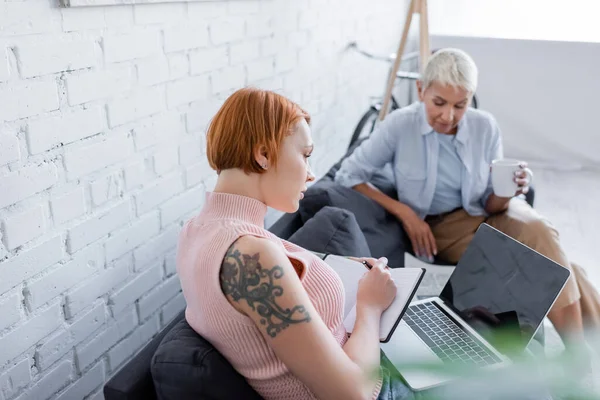 Tattooed Woman Writing Notebook Laptop Blank Screen Blurred Lesbian Girlfriend — Stock Photo, Image