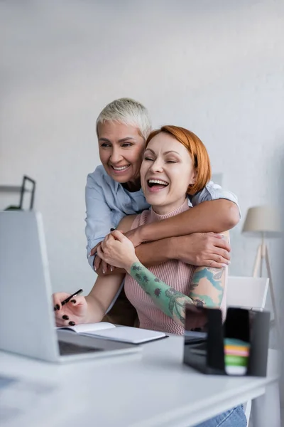 笑着的女同性恋者在家里抱着兴奋的女朋友 笑着接近模糊的笔记本电脑 — 图库照片