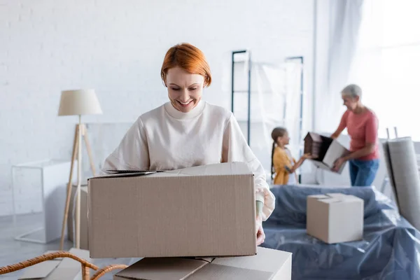 Smiling Lesbian Woman Holding Cardboard Package Blurred Family New Apartment — Stock Photo, Image