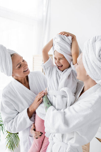 Happy lesbian women in bathrobes holding positive adopted daughter in bathroom 