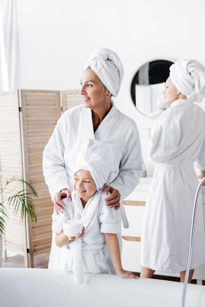 Woman in bathrobe hugging adopted daughter with cream in bathroom 