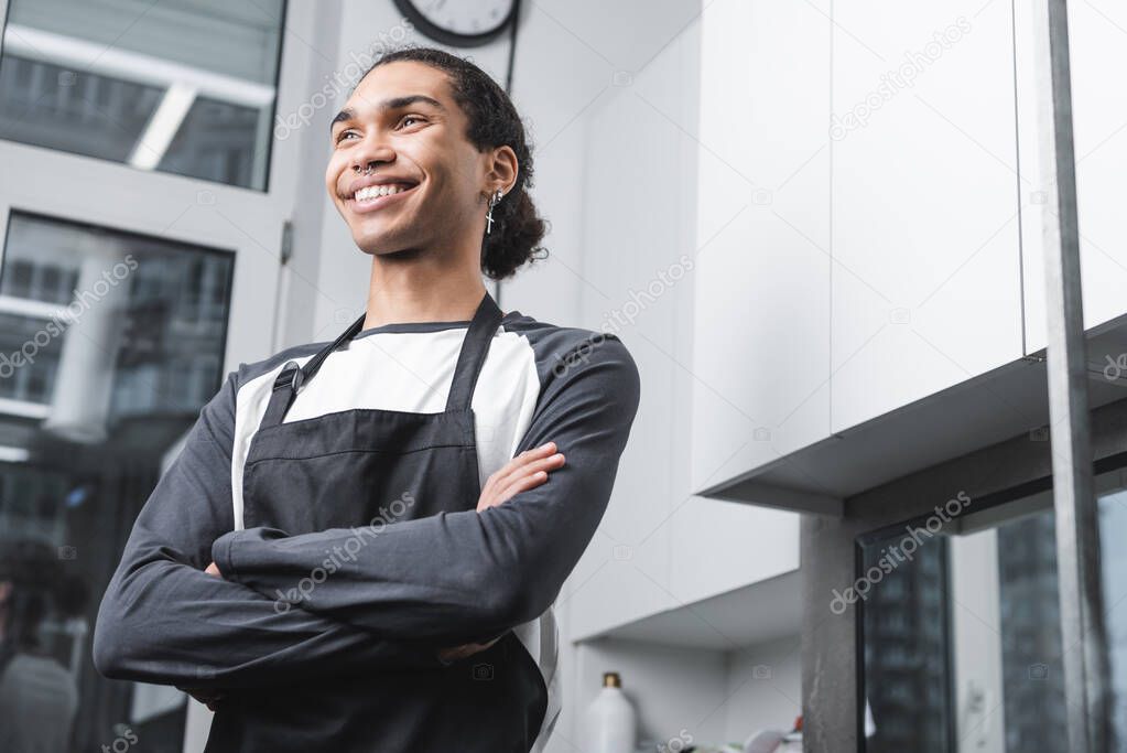 low angle view of happy african american barber standing with crossed arms in grooming salon