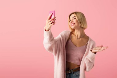 Yumuşak hırkalı mutlu genç bir kadın pembe üzerine izole edilmiş selfie çekiyor. 