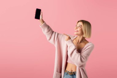 happy young woman in soft cardigan pointing at smartphone with blank screen isolated on pink  clipart