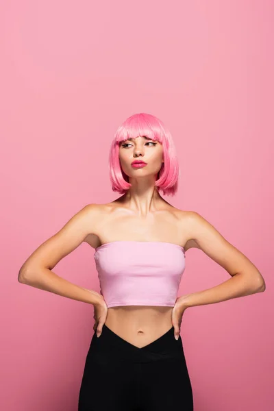 young model with bangs and colored hair posing with hands on hips isolated on pink