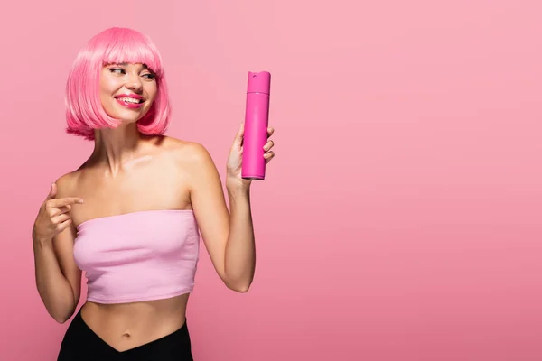 cheerful woman with colored hair pointing at bottle with spray isolated on pink