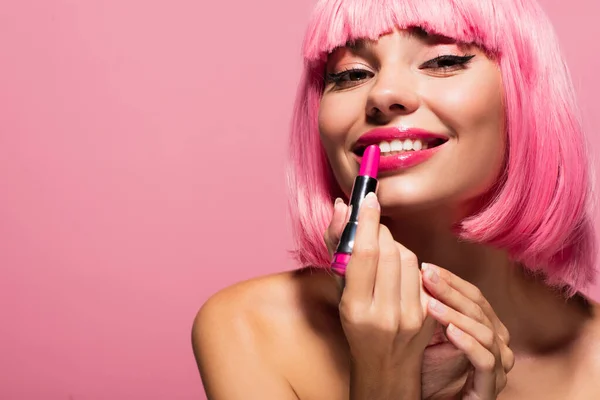 happy young woman with colored hair and bare shoulder applying lipstick isolated on pink