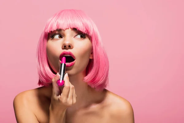 young woman with colored hair and bare shoulders applying lipstick and looking away isolated on pink