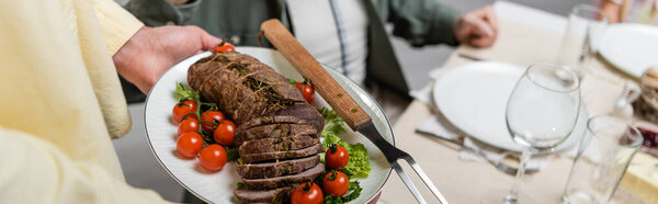 обрезанный вид человека, держащего тарелку с мясом и свежими овощами возле размытой семьи, баннер
