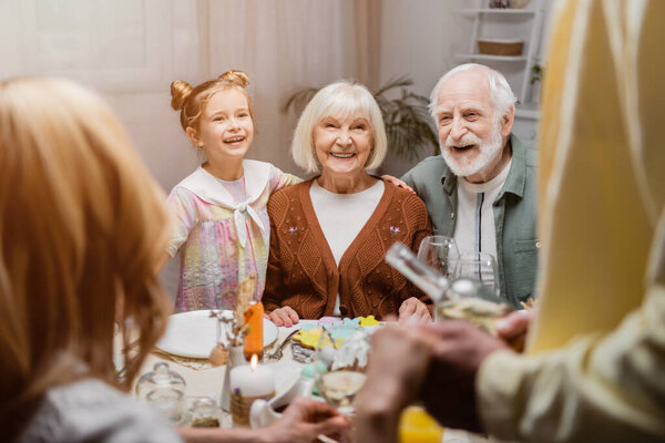 cheerful senior couple smiling near granddaughter and family on blurred foreground