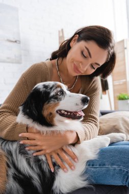Gülümseyen genç kadın koltukta otururken Avustralyalı çoban köpeğine sarılıyor.