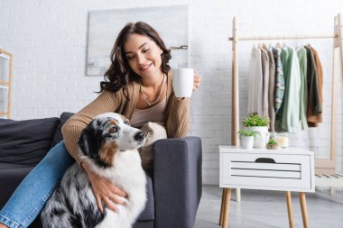 Avustralyalı çoban köpeğini kucaklayan ve elinde kahve tutan neşeli genç bir kadın.