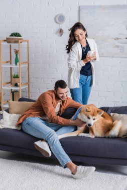 Kanepede oturan mutlu bir adam ve yanında bardakla Akia Inu 'nun köpeğine sarılıyor.