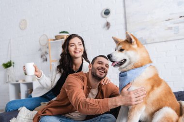pleased man sitting on couch and cuddling akita inu dog near happy girlfriend with cup clipart