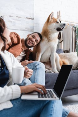 Gülümseyen genç kadın dizüstü bilgisayar kullanıyor mutlu erkek arkadaşının yanında Akia Inu köpeğine yaslanıyor.