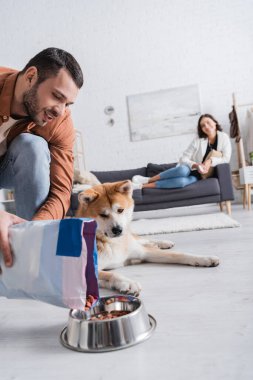 Sakallı adam Akita Inu köpeğinin yanındaki kaseye hayvan maması ekliyor.