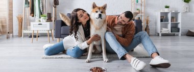 Genç ve mutlu bir çift oturma odasında evcil hayvan yiyeceğiyle akita inu dog 'a sarılıyor.