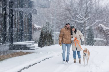 Sevgilisi ve köpeğiyle kar altında yürürken tasmasını tutan mutlu bir kadın.
