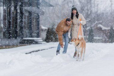Sevgilisi ve Akia Inu köpeğiyle kar altında yürürken tasmasını tutan neşeli kadın.