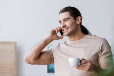 Uzun saçlı, elinde kahve fincanı olan ve akıllı telefonla konuşan mutlu bir adam. 