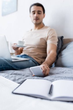 Yatak odasındaki akıllı telefona uzanırken elinde kahve fincanı olan bulanık bir adam.