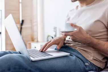 Yatak odasında dizüstü bilgisayar kullanırken elinde bir fincan kahve tutan adam görüntüsü.
