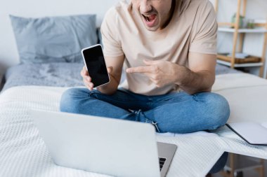 Kesik ağızlı bir adam yatak odasındaki video görüşmesi sırasında boş ekranla akıllı telefonu işaret ediyor. 