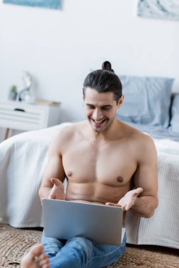 Uzun saçlı mutlu adam yatağın yanında oturuyor ve laptop kullanıyor.