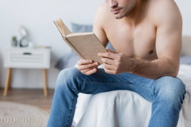 Yatakta oturmuş kitap okuyan üstsüz bir adam manzarası.