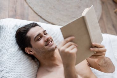 Yatakta uzanmış kitap okuyan mutlu ve üstsüz bir adam manzarası.