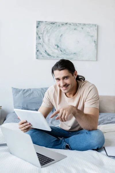 Radostný Muž Sluchátku Ukazuje Digitální Tablet Poblíž Notebooku Smartphonu Posteli — Stock fotografie