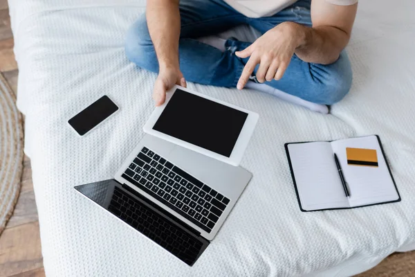 ベッドの上でノートパソコンやスマートフォンの近くに空白の画面を持つデジタルタブレットを指している人の — ストック写真
