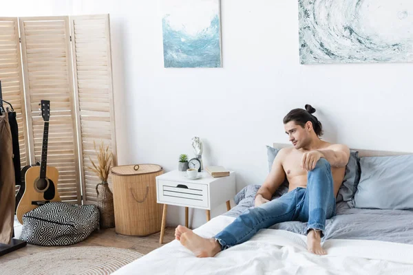 barefoot man in jeans resting on bed at home