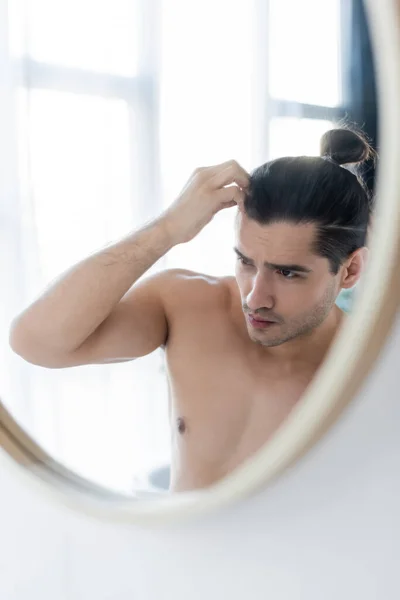 shirtless man adjusting hair bun and looking at mirror