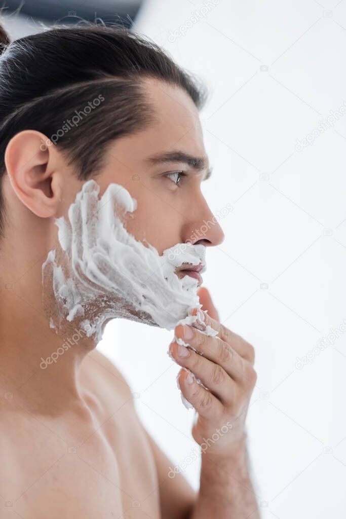 side view of shirtless man applying shaving foam on face in bathroom 