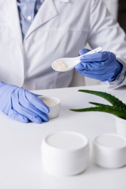 Lateks eldivenli kimyagerin elinde kremalı kaşık ve kase ile aloe yapraklarının yanında.