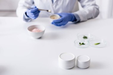 containers with cream near blurred laboratory assistant in latex gloves  clipart