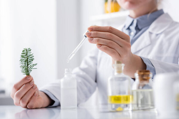 cropped view of laboratory assistant holding fir branch and pipette with serum near bottles in lab