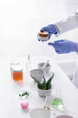 Laboratuvar asistanının lateks eldivenli, aloe ve kozmetik malzemelerinin yanında kremalı konteynır tutarken kısmi görüntüsü