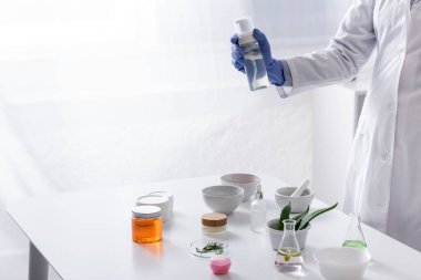 Laboratuvar malzemelerinin yanında plastik eldiven ve kozmetik ürünlerle dolu bir şişe tutan laboratuvar asistanının kısmi görüntüsü
