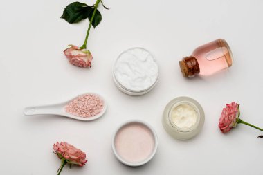 Pembe deniz tuzu yakınındaki çiçeklerin üst manzarası, gül suyu şişesi ve beyaz kremalı kaplar.