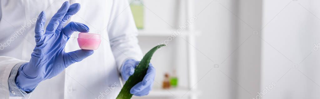 cropped view of laboratory assistant in latex gloves holding aloe leaf and container with cream, banner